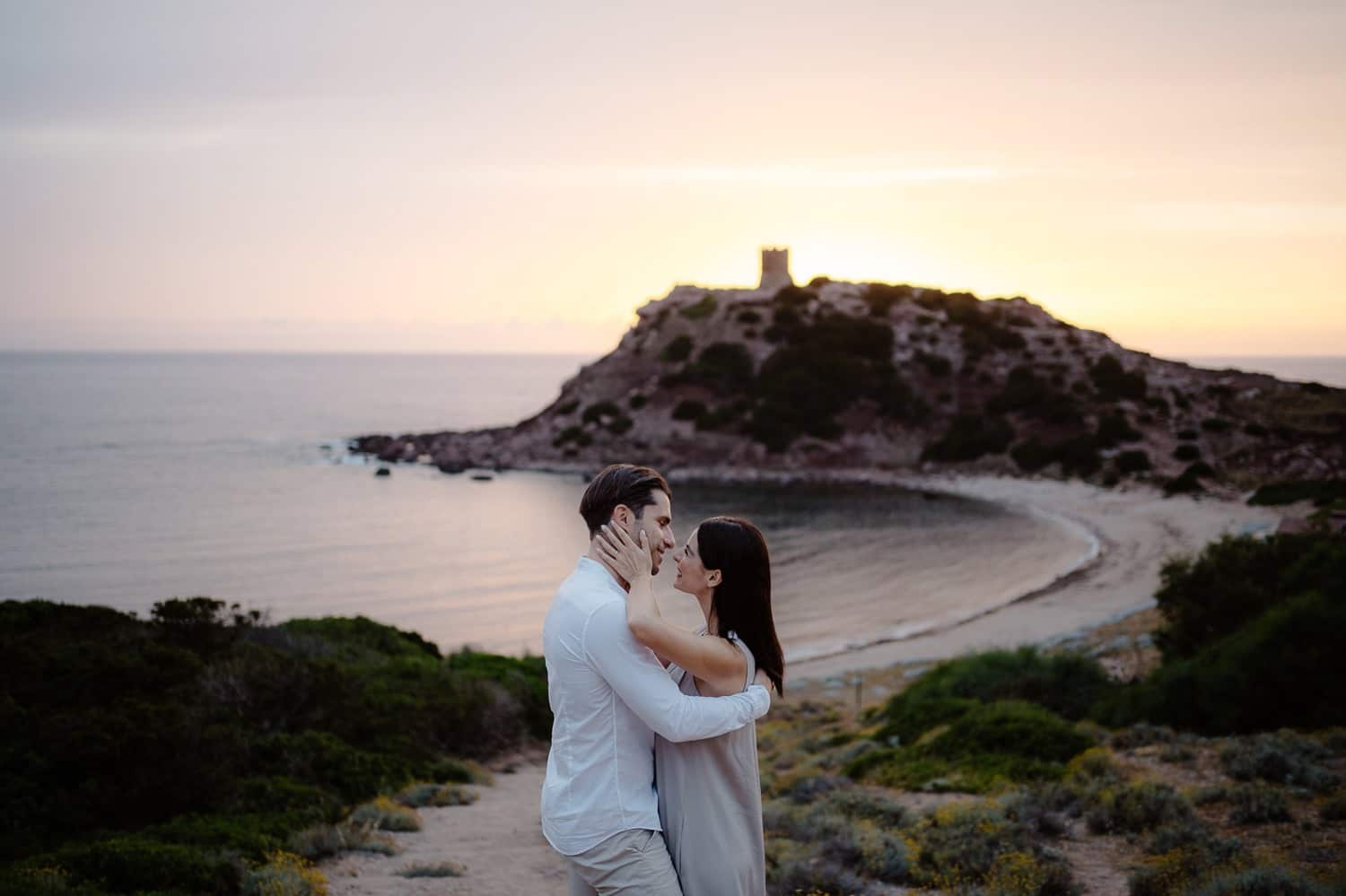 Couple kissing while sitting on a rock with the sea behind them, during their engagement in Alghero.