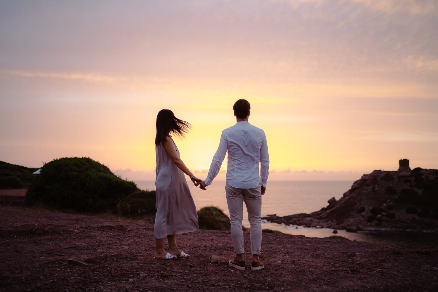 Couple walking on a beach and holding hands during their engagement in Alghero. The sea and coastline are in the background.