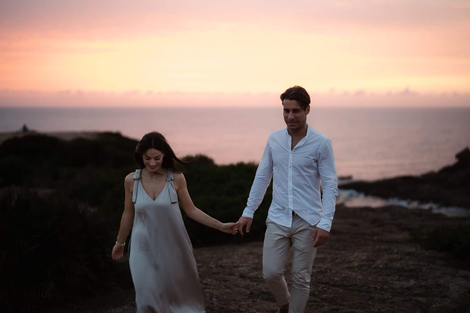Alghero photographer - A couple standing on a rocky cliff overlooking the ocean with the sun setting behind them, embracing each other