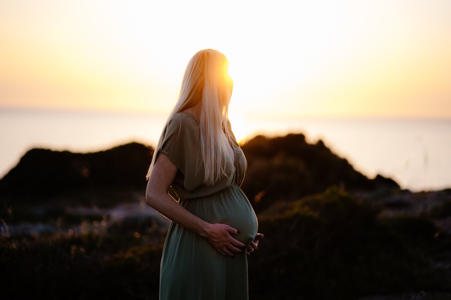 A beautiful maternity photo taken in the charming town of Alghero. The mother-to-be is standing in front of a colorful building with her hands on her belly, smiling at the camera.