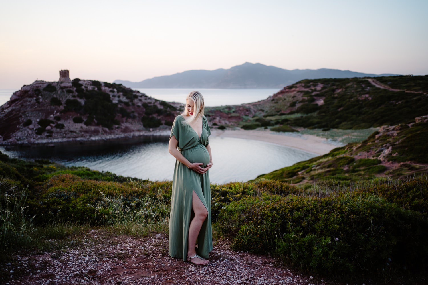 A heartwarming maternity photo taken in the picturesque town of Alghero. The mother-to-be is sitting on a stone staircase, looking down at her belly, with her husband standing next to her, holding her hand and looking at her lovingly.
